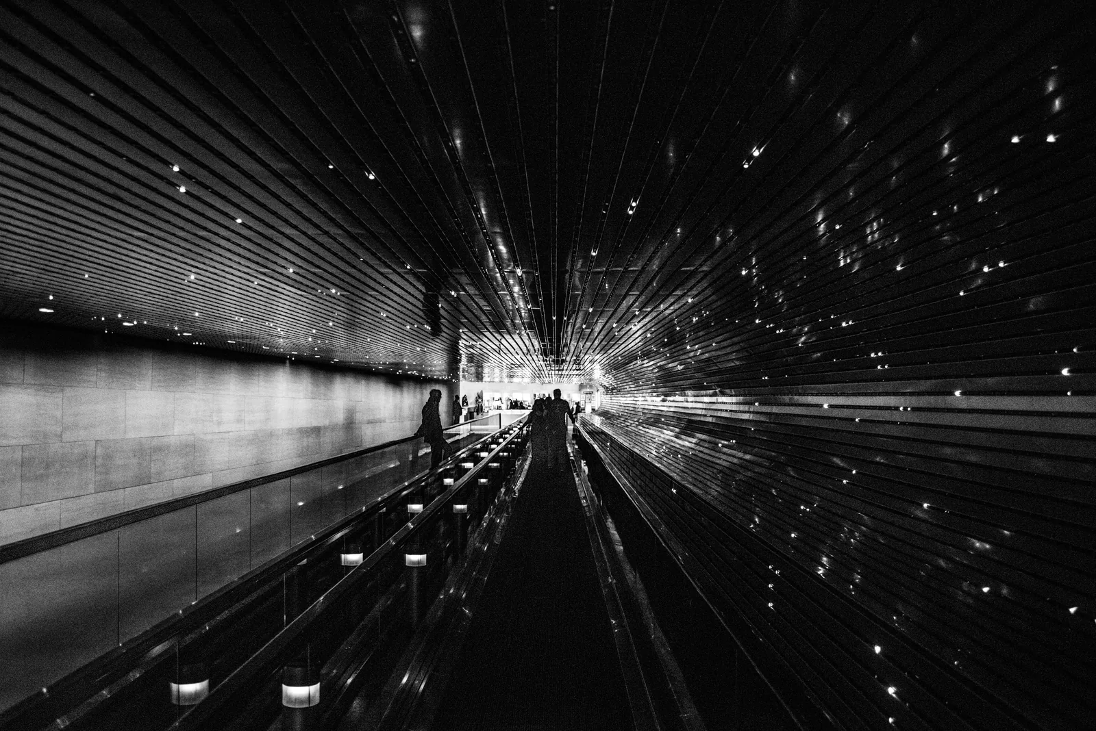 Pessoas andando numa esteira rolante de aeroporto ou estação, em foto preto e branco.