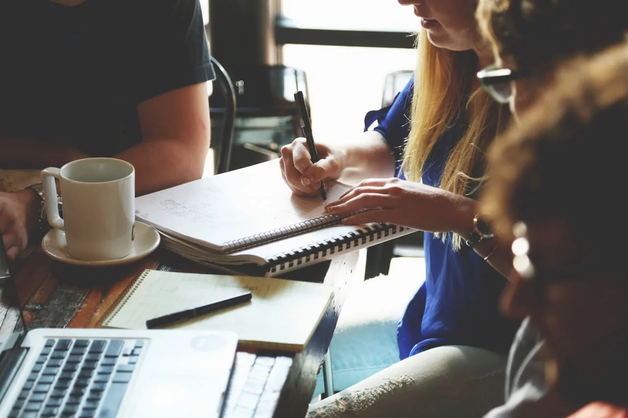 Três pessoas numa reunião em torno de uma mesa, com xícaras de café e um notebook.